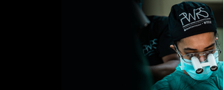 Un chirurgien dans une salle d'operation, portant une coiffe chirurgicale et un masque facial, regarde concentre devant lui avec un masque offrant un grossissement.