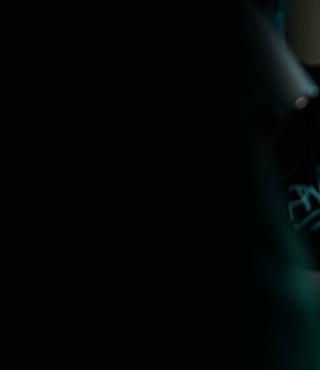 Texte alternatif : Un chirurgien dans une salle d'operation porte une coiffe avec le logo "PWRS" et un masque facial. Il utilise une visiere speciale avec des lentilles grossissantes et est concentre sur une intervention medicale.