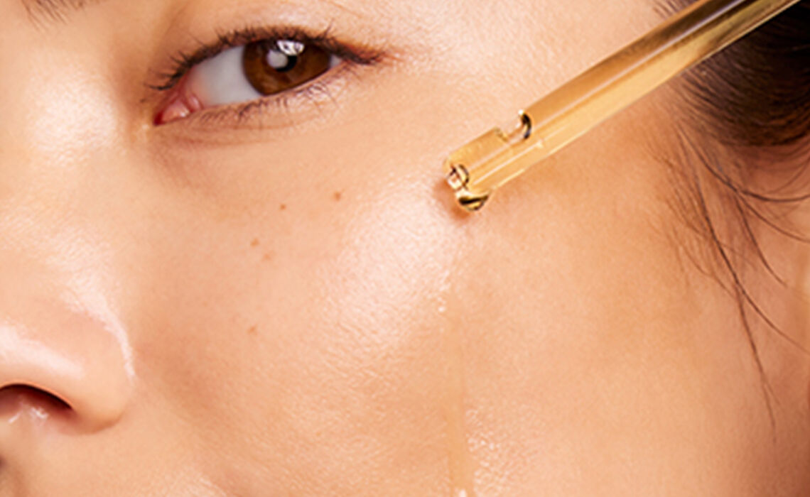 Een close-up van een gezicht van een vrouw met een parfumflesje dat tegen de huid druppelt, waardoor een glanzende uitstraling zichtbaar is.