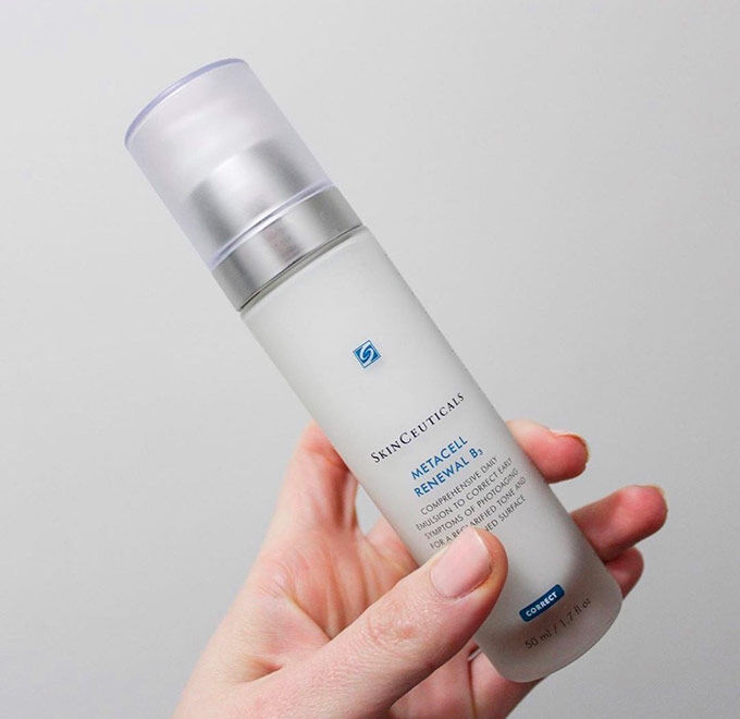 Een hand houdt een flesje van Skinceuticals Metacell Renewal B, een gezichtserum met een blauwe en zilveren dop.