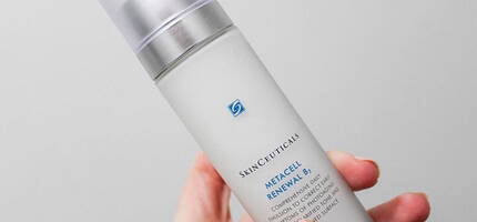 Een hand houdt een flesje van Skinceuticals Metacell Renewal B, een gezichtserum met een blauwe en zilveren dop.