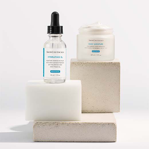 A skincare product display featuring a dropper bottle of Hydrating B5 serum and a jar of Daily Moisture cream, both on neutral-toned blocks.