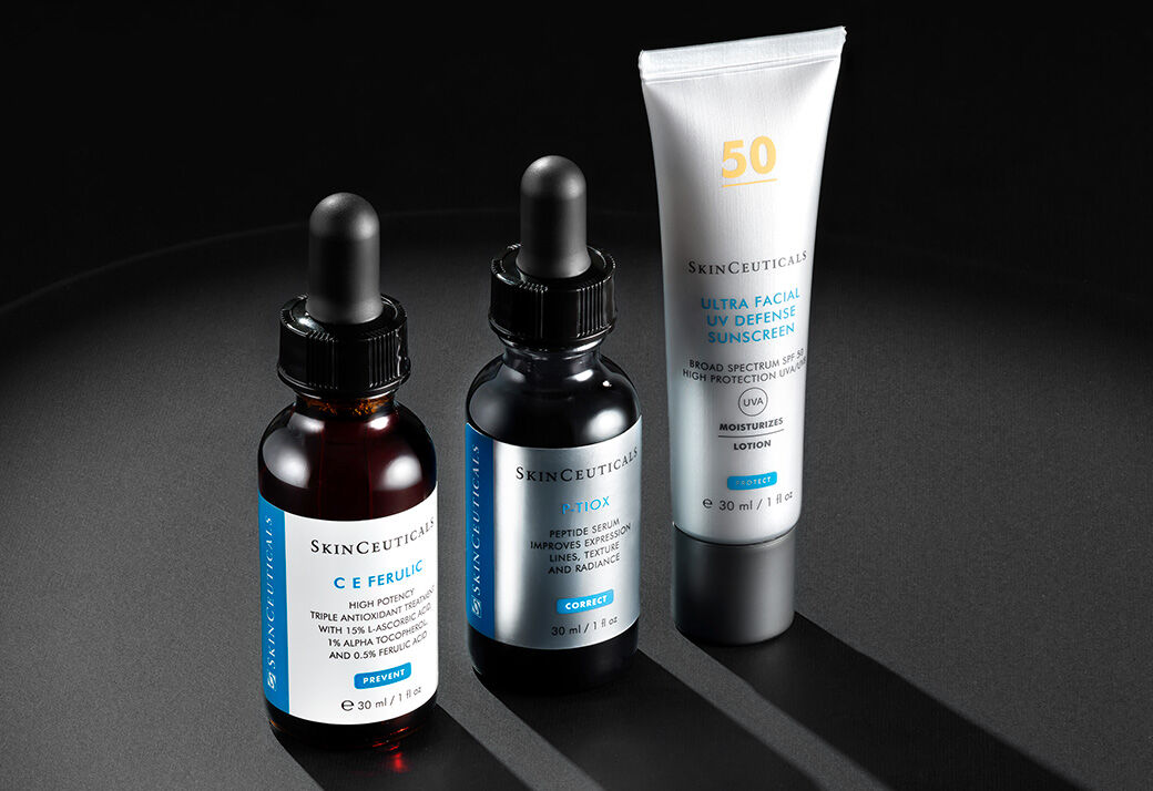 Drie flessen huidverzorgingsproducten van SkinCeuticals op een donkere achtergrond: een serum met pipet, een antioxiderend serum en een zonnebrandcreme.