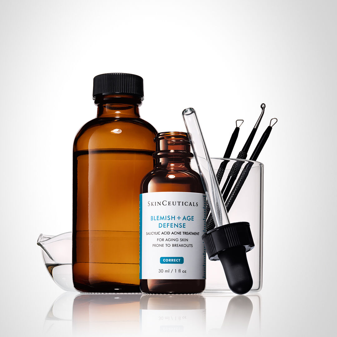 Een set huidverzorgingsproducten, waaronder een amberkleurige fles, een serumfles van SkinCeuticals met een etiket 'Blemish + Age Defense', een pipet en enkele huidverzorgingsinstrumenten in een laboratoriummateriaal.
