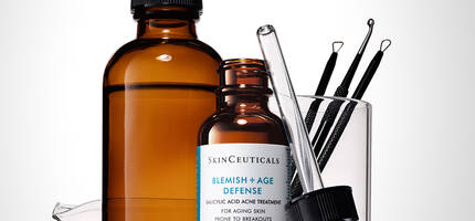 Een set huidverzorgingsproducten, waaronder een amberkleurige fles, een serumfles van SkinCeuticals met een etiket 'Blemish + Age Defense', een pipet en enkele huidverzorgingsinstrumenten in een laboratoriummateriaal.