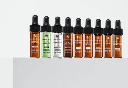 Een reeks van negen glazen pipetflessen met huidverzorgingsserums in verschillende kleuren, uitgestald op een witte ondergrond.