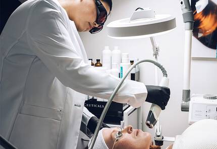 Een huidtherapeut voert een behandeling uit op een patient in een schoonheidssalon, terwijl de patient op een behandeltafel ligt.